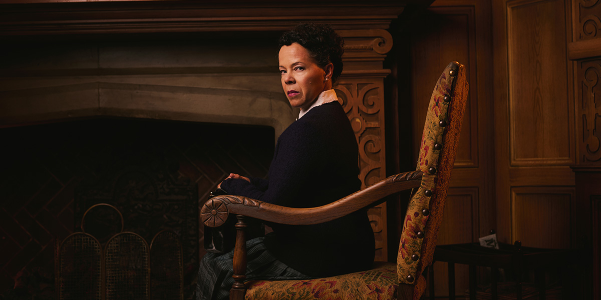 A woman with curly hair sits in a patterned chair, gazing intently, with a fireplace in the background.