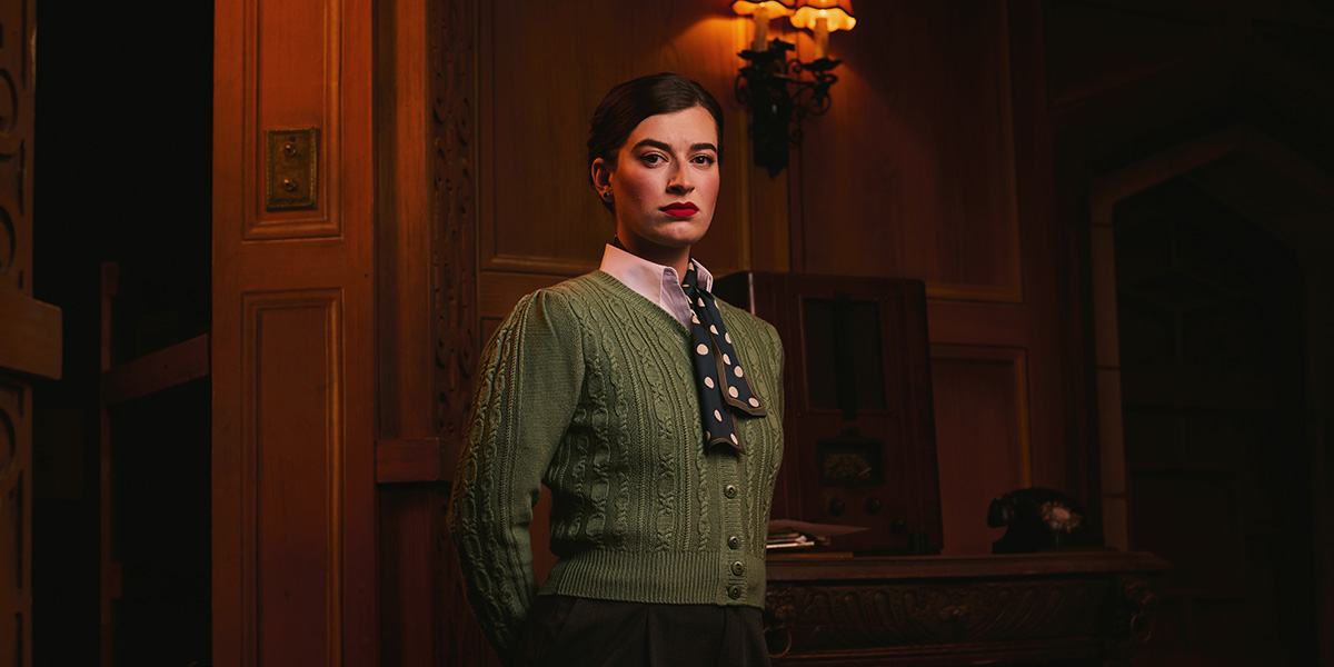 A woman in a green cable-knit sweater and polka dot tie stands confidently in a wooden interior.