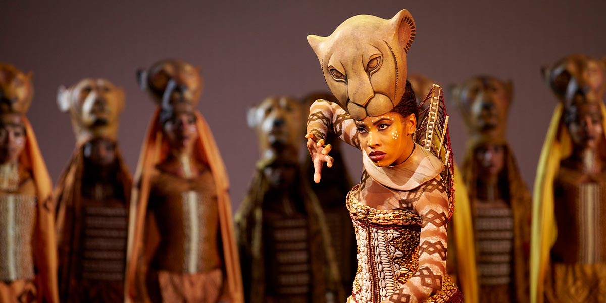 Daniela Pobega as Nala, wearing a lion costume, stands on stage with dramatic lighting in the background.