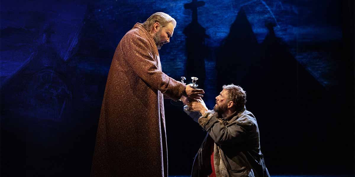 Two actors in period costumes exchange a silver candlestick on a dark stage with blue lighting.