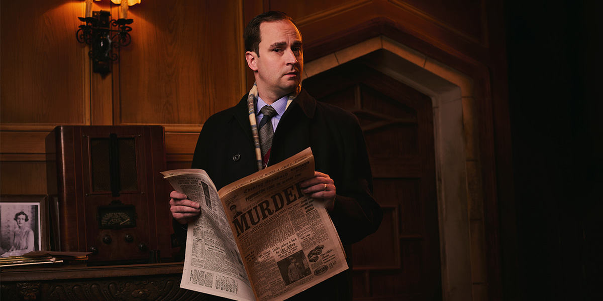 A man in a black coat holds a newspaper titled "MURDER" in a dimly lit, wooden room.
