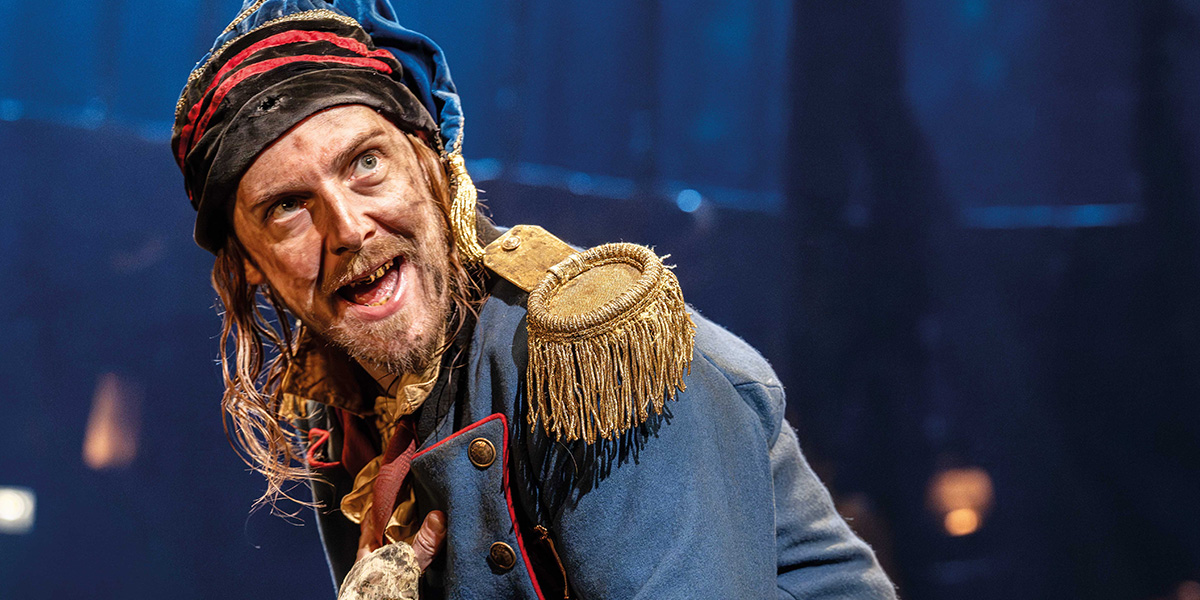 Harry Hepple as Thenardier, wearing a dark coat and hat, stands with a mischievous expression on stage.