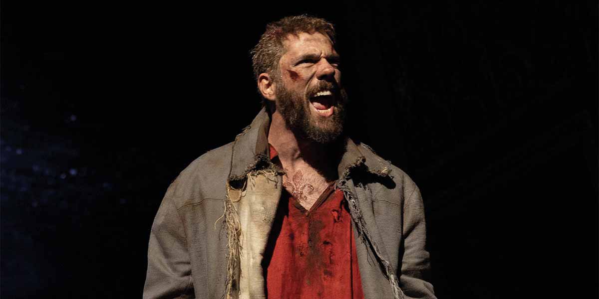 Ian McIntosh as Valjean, shouting passionately, wearing a tattered grey coat over a red shirt.