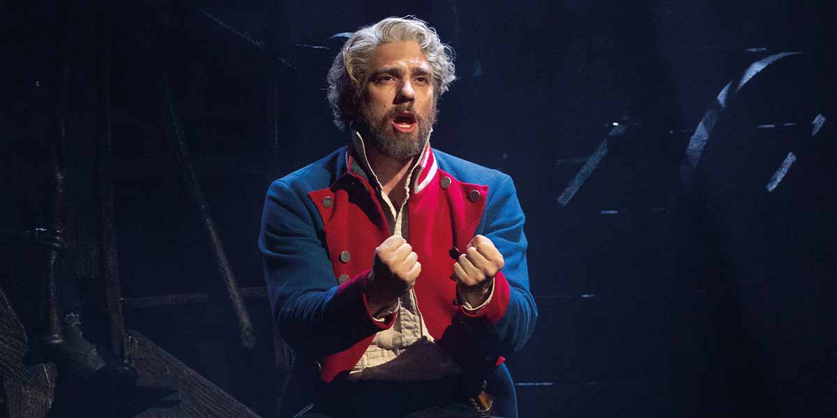 Ian McIntosh as Jean Valjean passionately performs, wearing a blue and red coat, against a dark background.