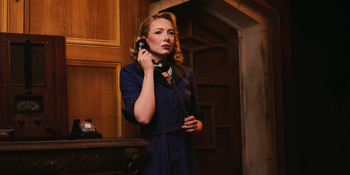Mollie stands by a vintage telephone, holding the receiver, with a serious expression against a wooden backdrop.