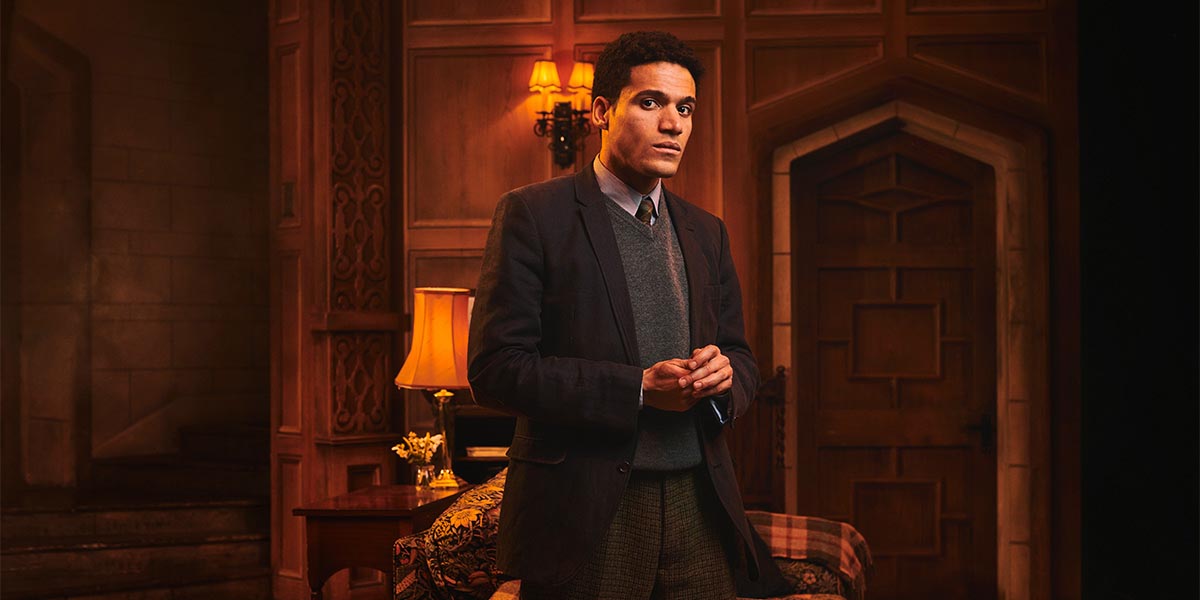 A man stands thoughtfully in a dimly lit room, wearing a dark blazer and grey sweater.