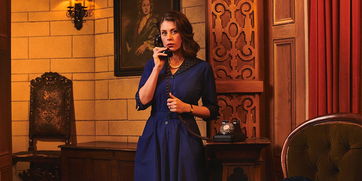 A scene from The Mousetrap featuring actors in period costumes on stage, with dramatic lighting and a wooden set.