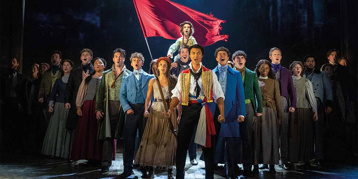 A group of performers passionately sings, holding a red flag, dressed in period costumes from Les Misérables.