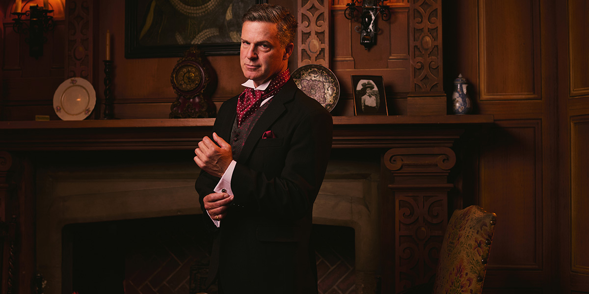 A man in formal attire adjusts his cufflinks, standing confidently in front of a wooden fireplace.
