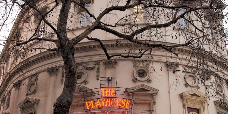 The Playhouse Theatre in London, featuring a red brick facade and a clear blue sky background.