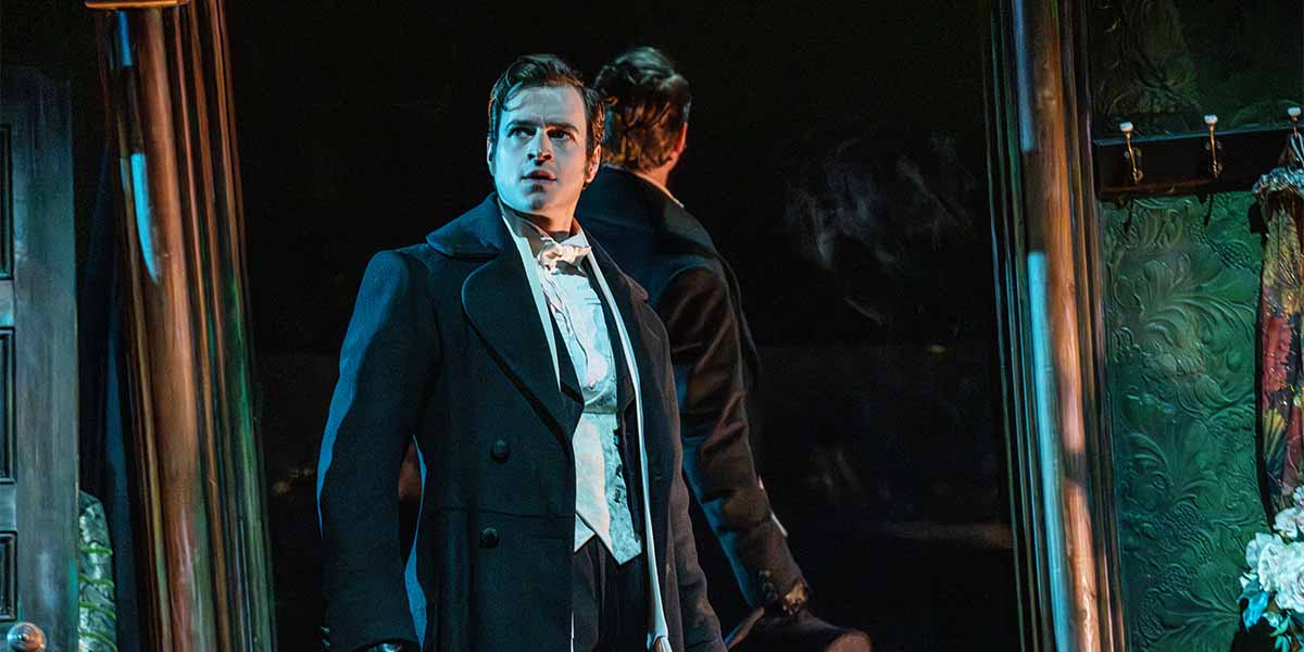 Ashley Gilmour as Raoul stands in a dramatic pose, wearing a black coat, against a dark background.