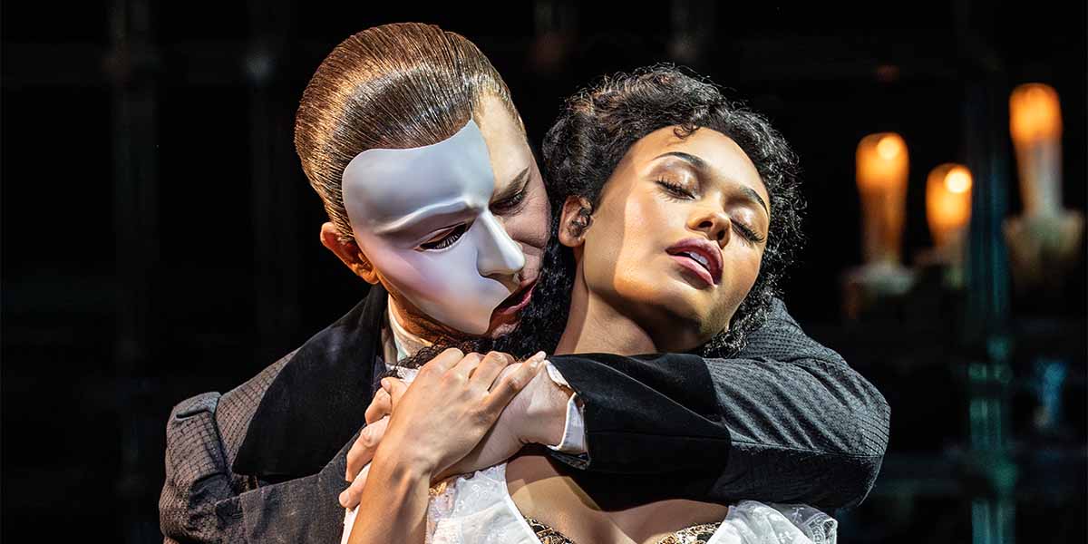 Dean Chisnall as The Phantom embraces Beatrice Penny-Touré as Christine Daaé, with candles in the background.