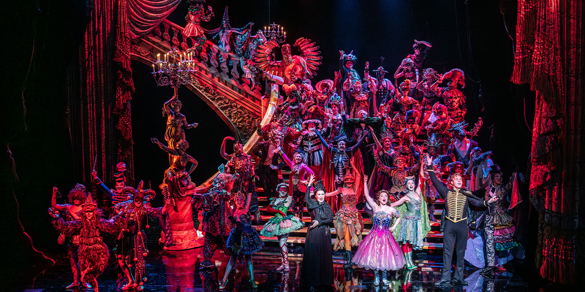 A vibrant ensemble in elaborate costumes performs on stage, featuring a grand staircase and dramatic lighting.