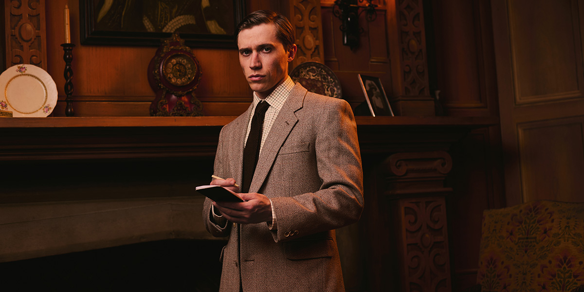 A man in a suit stands on stage, holding a script, with a spotlight illuminating him.