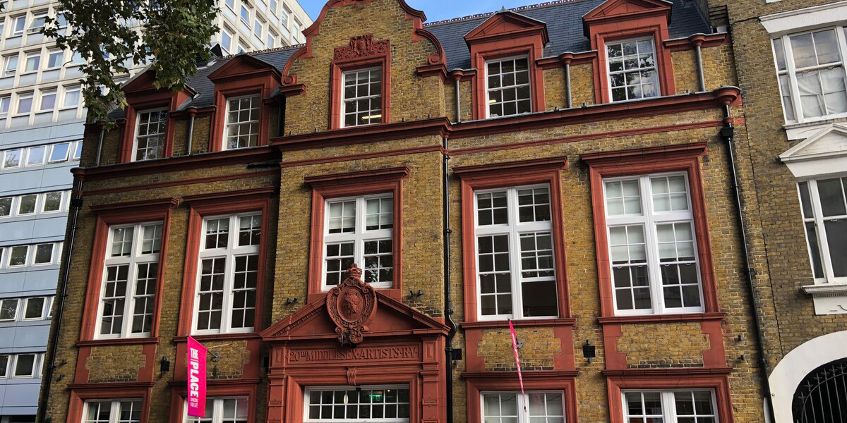 Red-brick building with white windows, featuring a decorative entrance and a pink banner in the foreground.