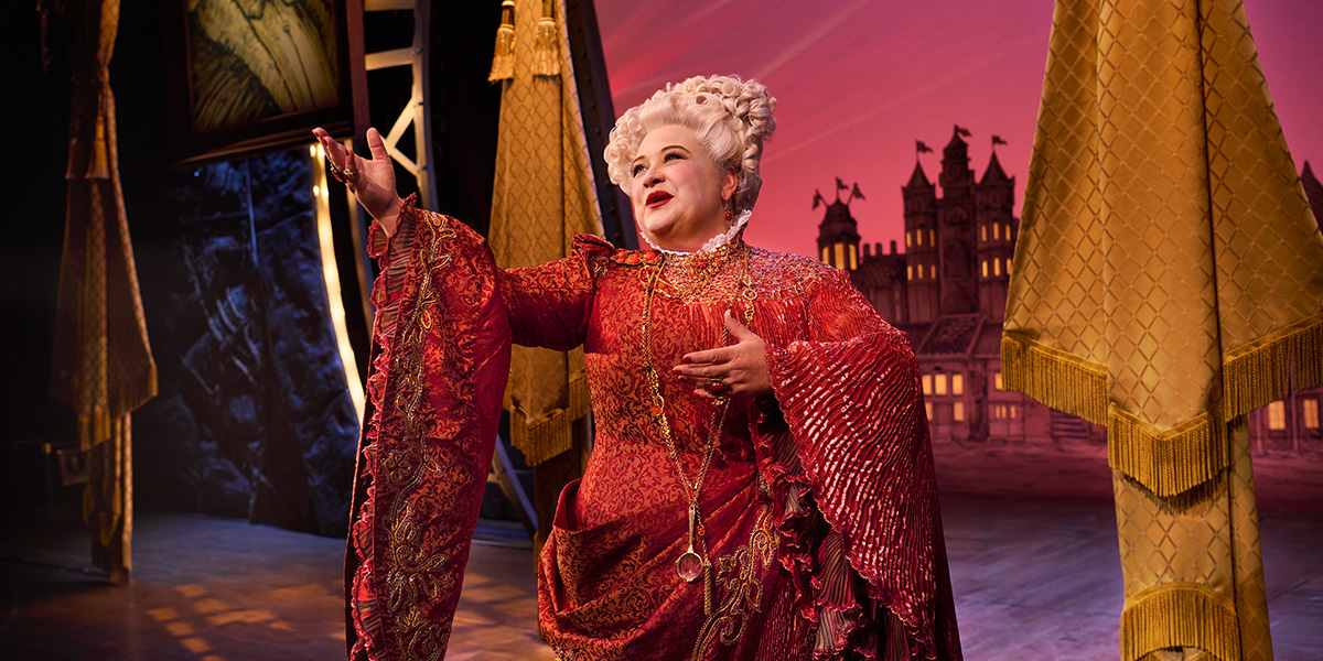 Claire Machin as Madame Morrible, wearing a dark costume with a textured cape, stands confidently on stage.