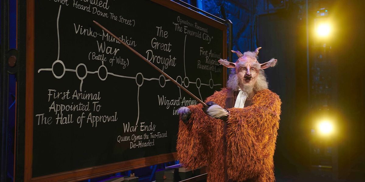 A green witch costume with a pointed hat, set against a dark stage background.