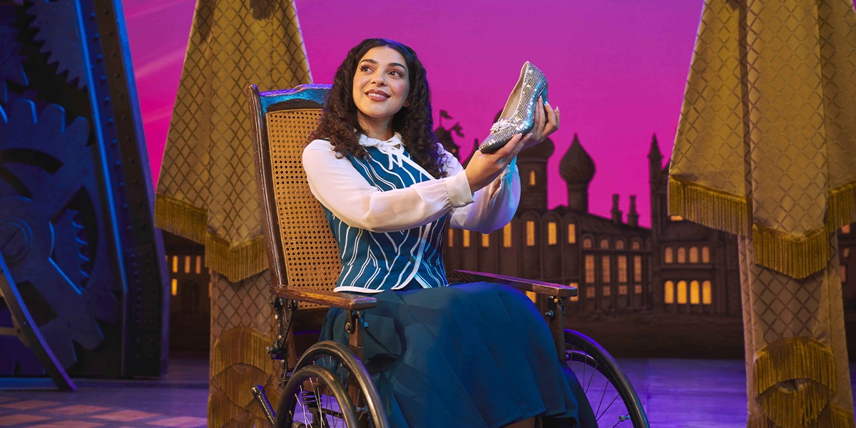 Hannah Qureshi in a green costume, smiling on stage with a dark backdrop and bright lights.