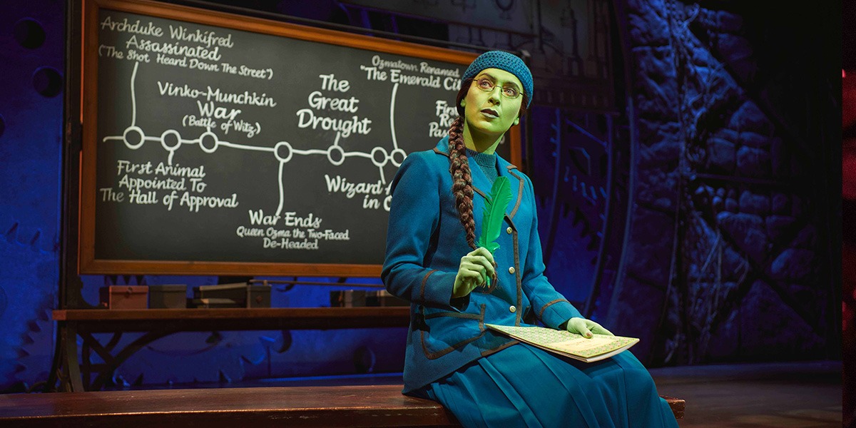 A woman in a green dress stands confidently on stage, with a dark curtain backdrop.