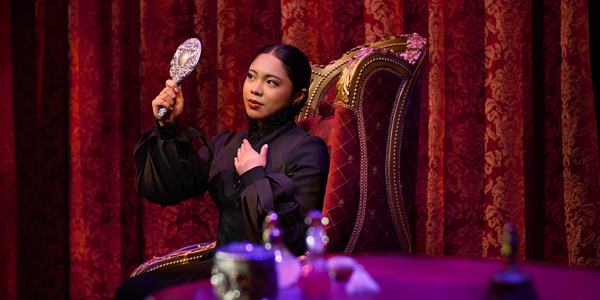 Yara Fabricante as Nessarose, wearing a green dress, stands on stage with a spotlight behind her.