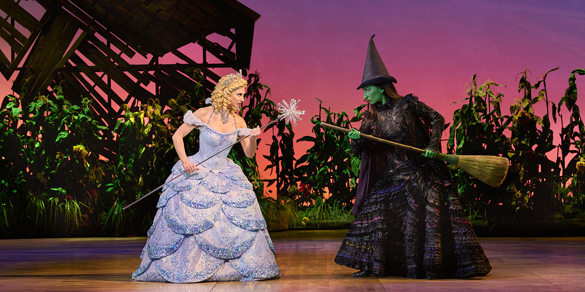 Zizi Strallen as Glinda and Emma Kingston as Elphaba pose together, wearing vibrant costumes on stage.