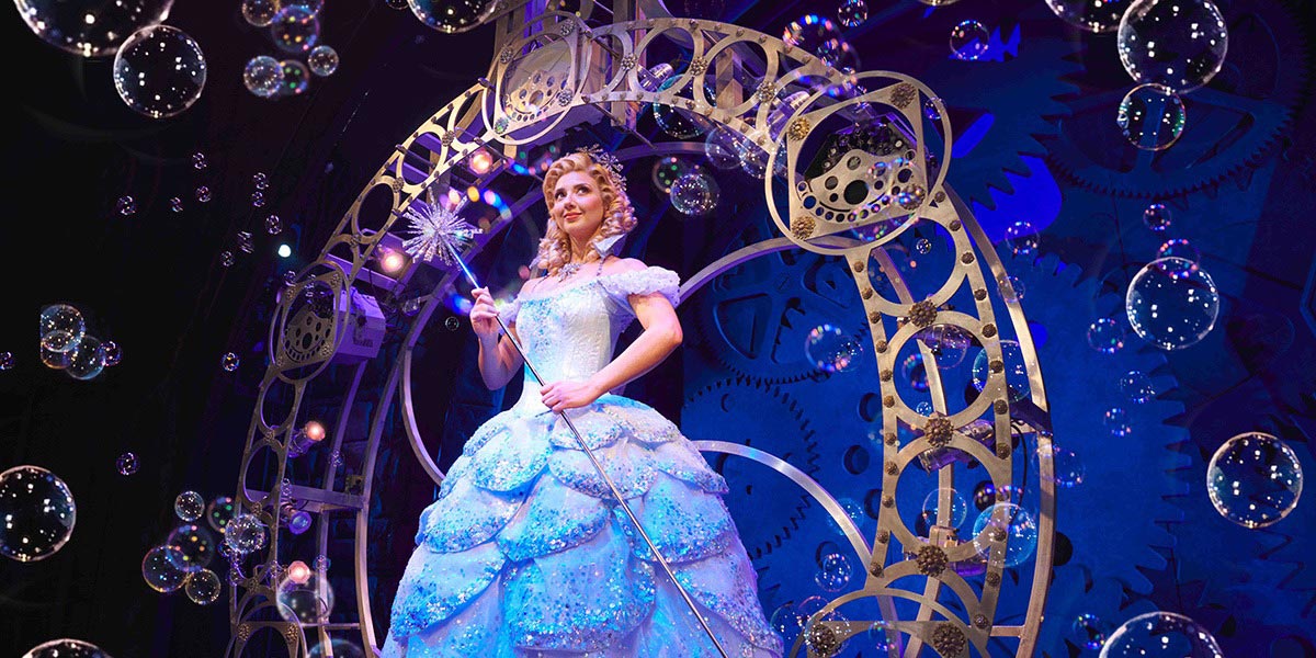 Zizi Strallen as Elphaba, wearing a green costume, stands on stage with dramatic lighting and a dark background.