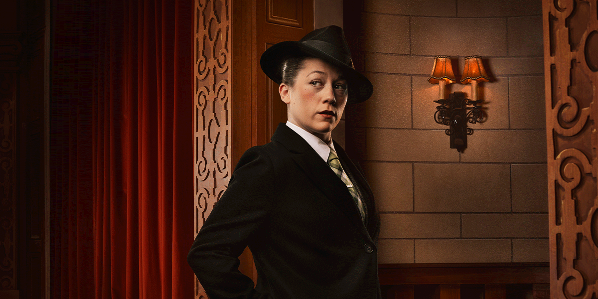 A woman in a black suit and hat stands confidently against a textured wall, with warm lighting.