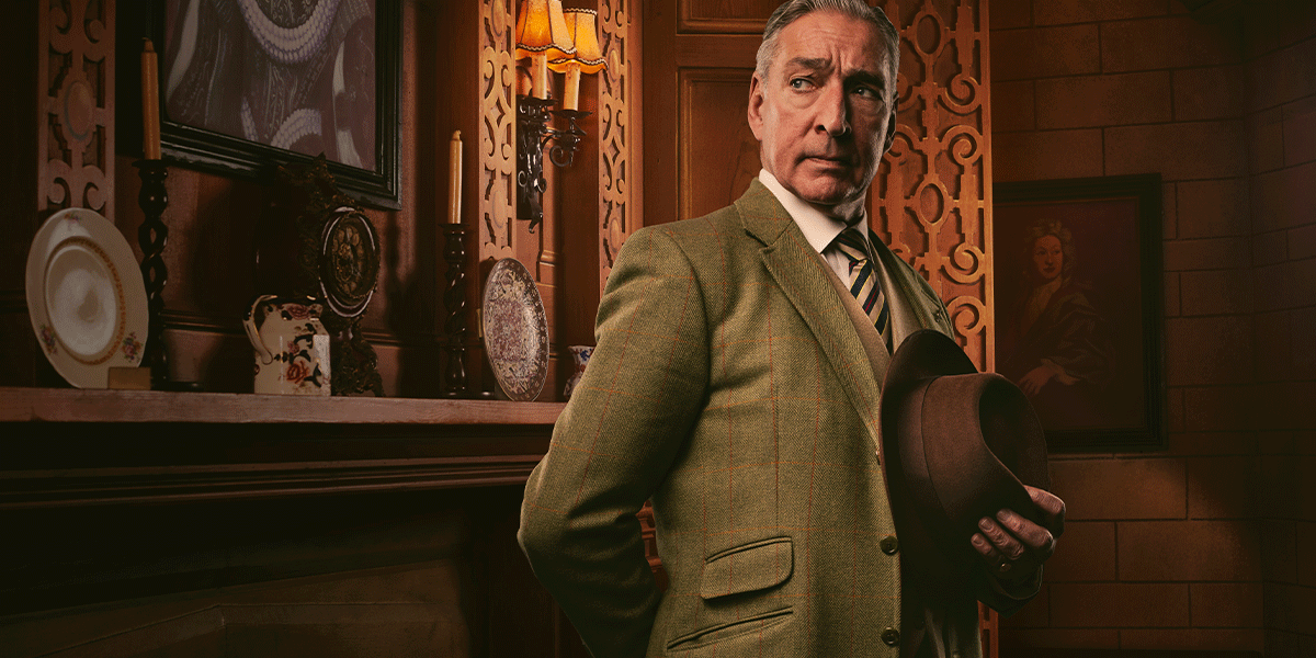 A man in a green tweed suit holds a brown hat, standing in a warmly lit room with decor.