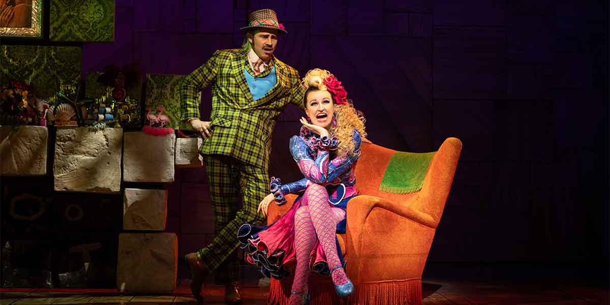 A person in a checked green suit stands against an orange armchair where a person with long, curly blonde hair wearing a brightly coloured dance costume is seated.