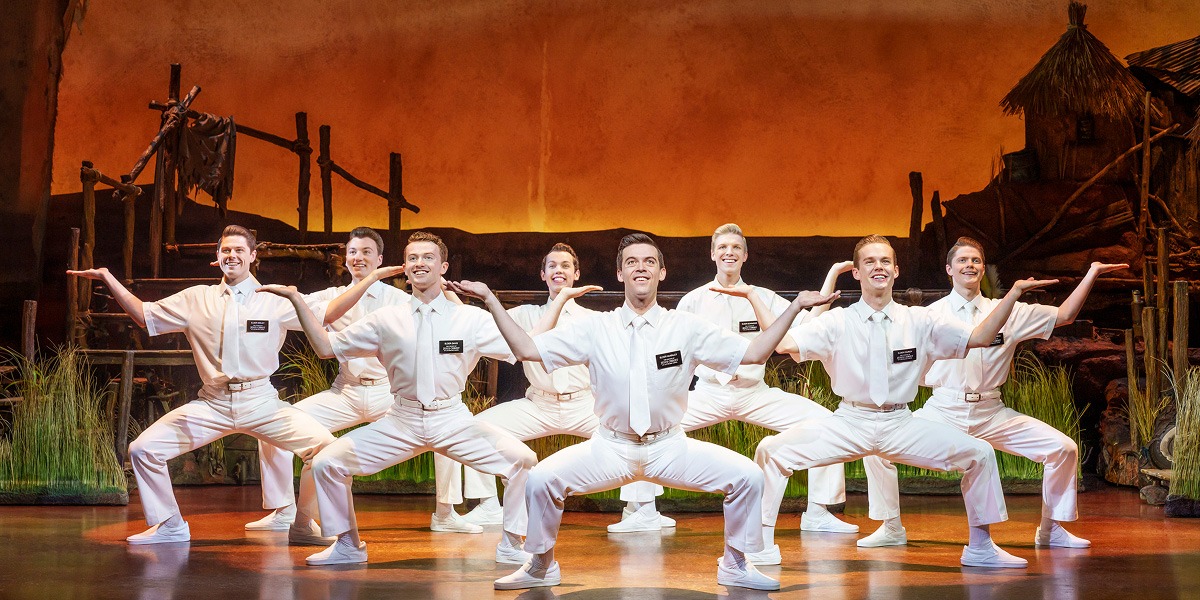 Eight performers in white costumes pose energetically on stage, with a vibrant sunset backdrop and greenery.