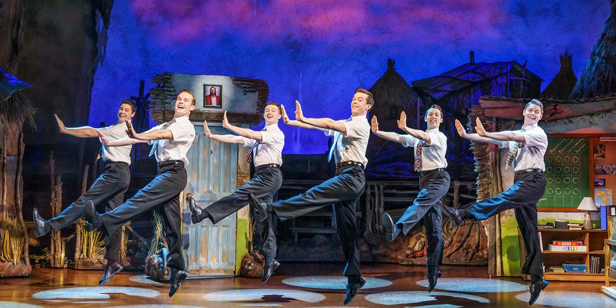 Six male performers in white shirts and black trousers dance energetically on stage against a colourful backdrop.