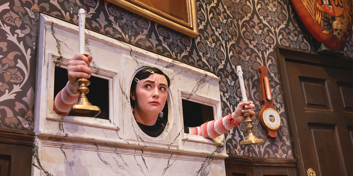 A woman with dark hair in a black dress stands on stage, holding a script, with dramatic lighting.