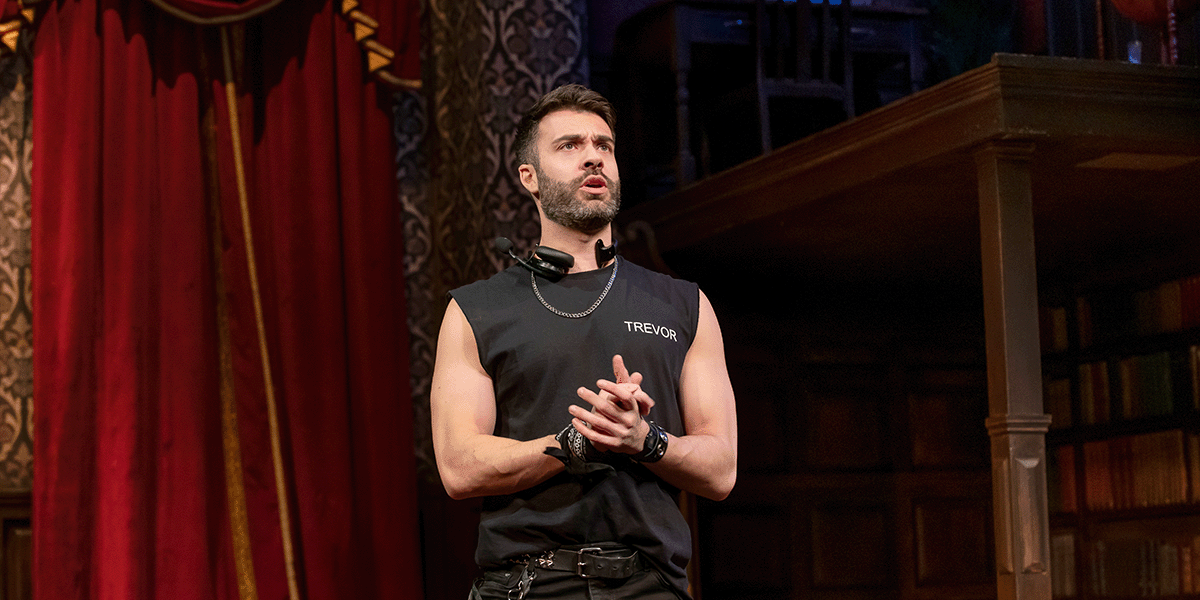 A man in a suit stands on stage, holding a script, with dramatic lighting and a dark backdrop.