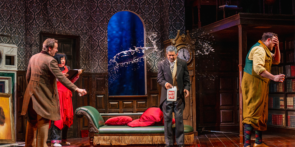 A chaotic scene from "The Play That Goes Wrong" featuring actors in mismatched costumes amidst scattered props. One person is covered in water.