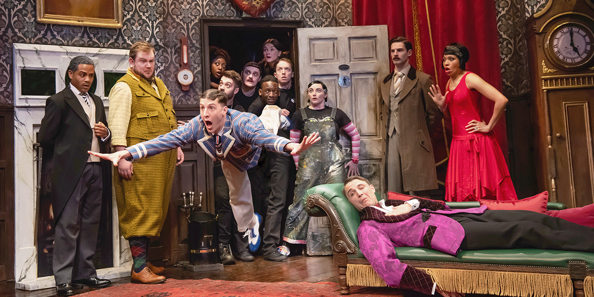 A group of actors in period costumes performs a comedic scene on a cluttered stage with props.