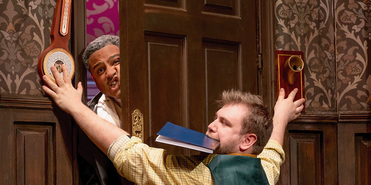 A man in a suit gestures animatedly on stage, with dramatic lighting and a theatrical backdrop.