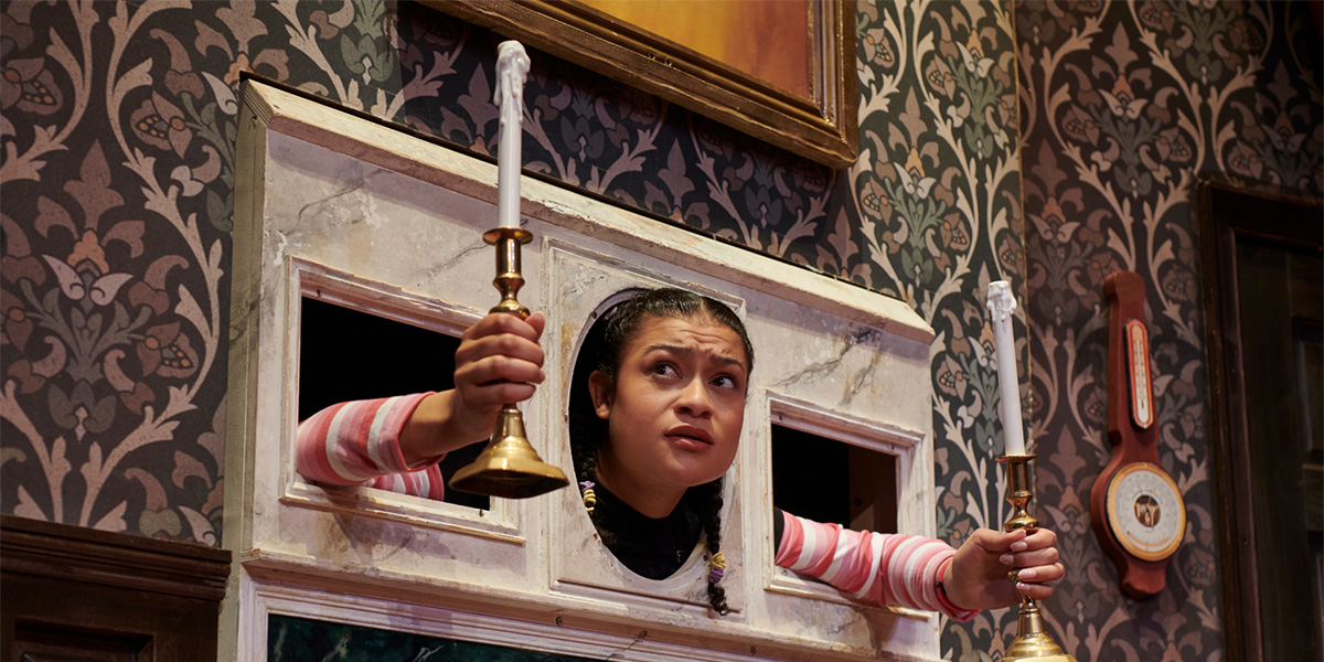 A person poke their head out of a hold in a fireplace and holds two candelbra's.