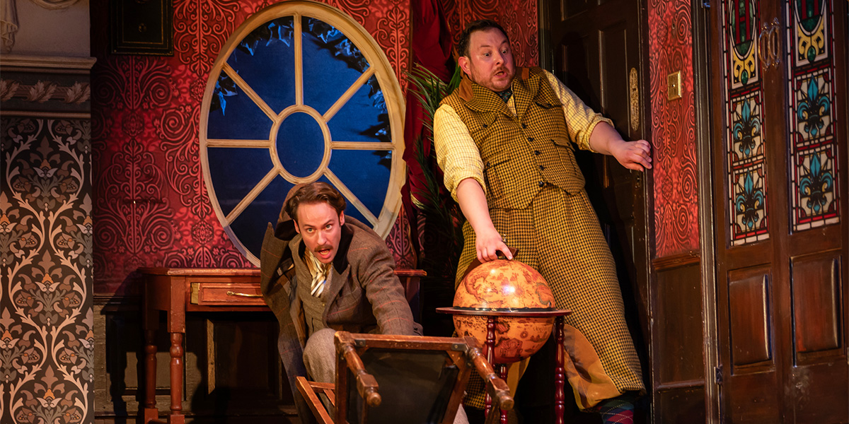 A group of actors in various styles of costume performs a comedic scene on a cluttered stage, with props scattered around.