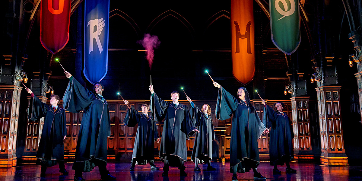 The cast stands together on stage, dressed in theatrical costumes, with dramatic lighting and a dark background.