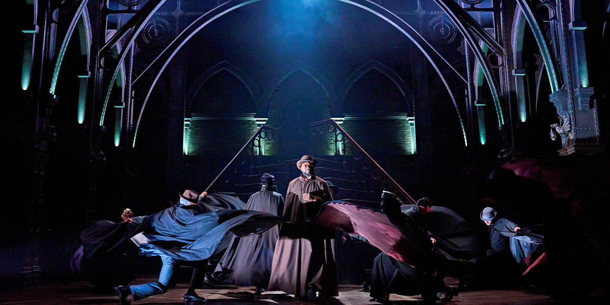 A diverse group of actors in costume poses together on stage, with dramatic lighting and a dark backdrop.