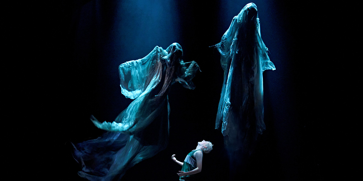 A performer in a green costume looks up at two ethereal, flowing figures in blue light.