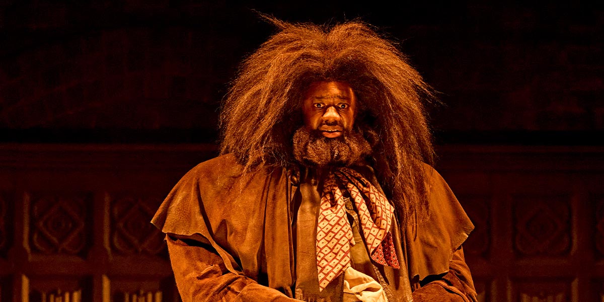 Gabriel Fleary as Hagrid, wearing a brown coat, stands in a forested area, surrounded by trees.