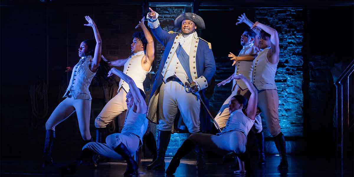 Akmed Junior Khemalai performs centre stage, surrounded by dancers in period costumes against a blue-lit backdrop.