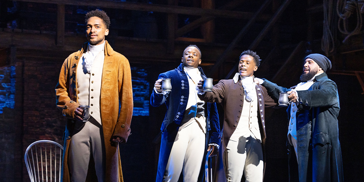 Five male performers in period costumes pose together, showcasing vibrant fabrics and expressive facial expressions.