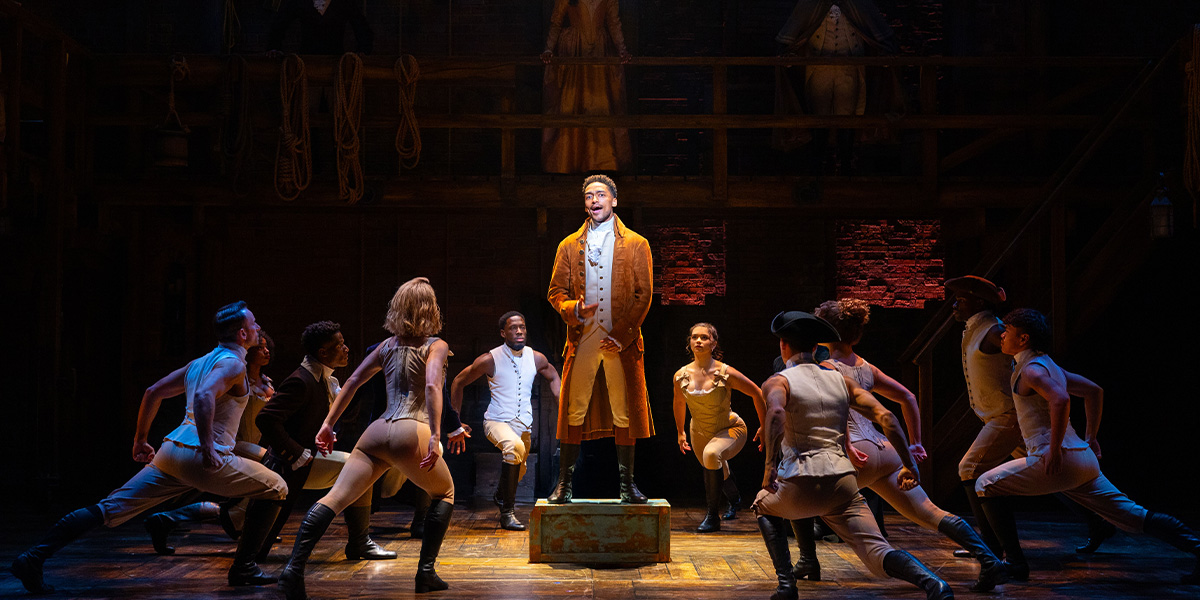 Alex Sawyer as Alexander Hamilton stands centre stage, surrounded by ensemble members in period costumes, dramatic lighting.