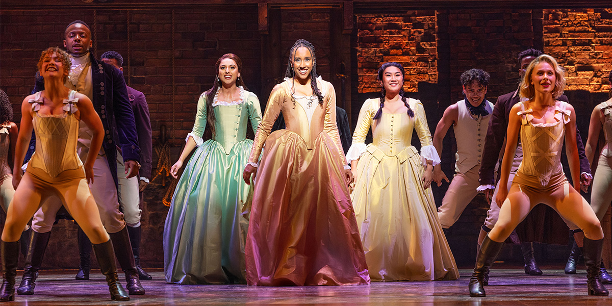 Five women in period costumes pose together, showcasing vibrant fabrics and intricate details against a neutral background.