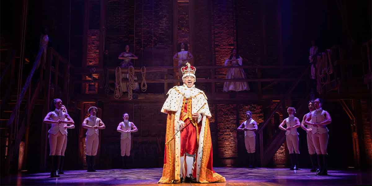 A performer in a royal costume stands centre stage, surrounded by dancers in neutral attire.
