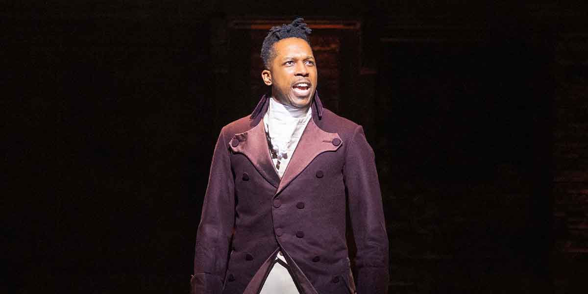 Leslie Odom Jr. performs passionately in a dark setting, wearing a burgundy coat and white shirt.