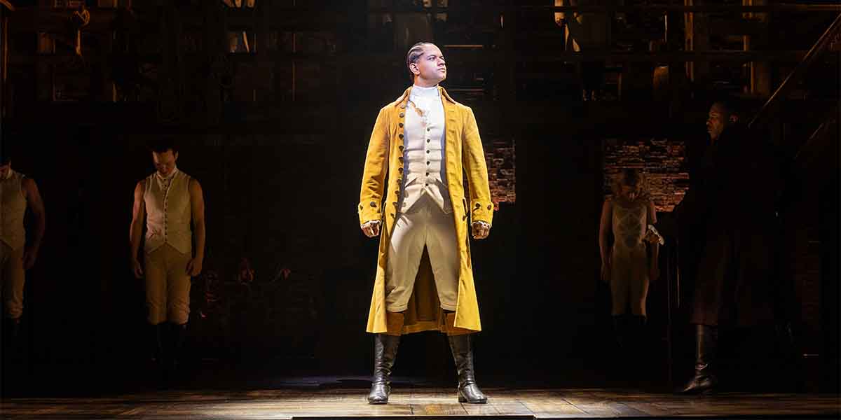 A performer in a yellow coat stands confidently centre stage, with dimly lit figures in the background.