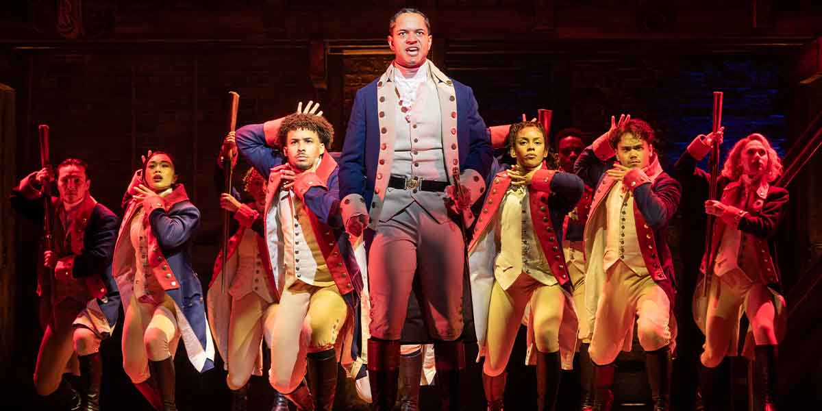 A group of performers in historical costumes, striking dynamic poses with props, illuminated by dramatic lighting.